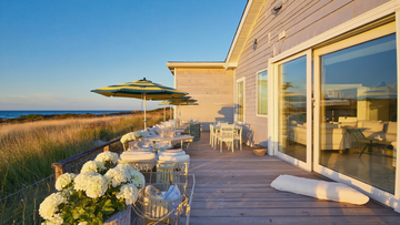 Luxury beachfront home deck with outdoor seating, striped umbrellas, and ocean views at sunset