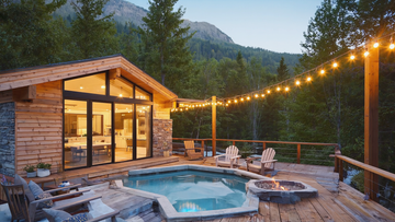 Modern mountain cabin with outdoor hot tub, fire pit and string lights surrounded by forest and scenic alpine views at dusk